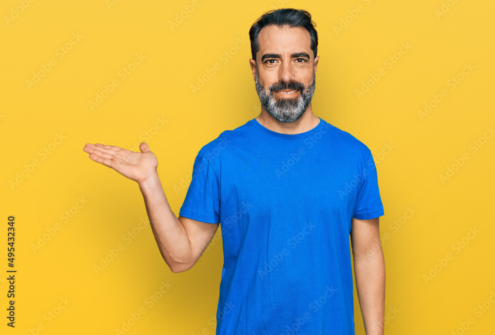 Middle aged man with beard wearing casual blue t shirt smiling cheerful presenting and pointing with palm of hand looking at the camera. Middle aged man with beard wearing casual blue t shirt smiling cheerful presenting and pointing with palm of hand looking at the camera.