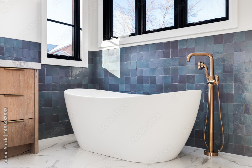A beautiful, luxurious bathroom detail shot of a white standalone tub and gold faucet sitting on white marble tiles and in front of a blue tiled wall. Stock Photo | Adobe Stock A Beautiful, Luxurious Bathroom Detail Shot Of A White Standalone Tub And Gold Faucet Sitting On White Marble Tiles And In Front Of A Blue Tiled Wall. Stock Photo | Adobe Stock