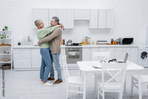 Side view of senior woman hugging husband near papers and devices in kitchen. © LIGHTFIELD STUDIOS