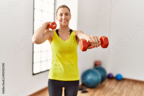 Young blonde girl smiling happy training using dumbbells at sport center.