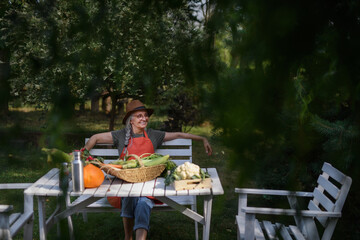 Happy senior farmer sitting at table and looking at harvest outdoors in garden.