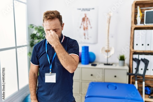 Middle age physiotherapist man working at pain recovery clinic tired rubbing nose and eyes feeling fatigue and headache. stress and frustration concept.