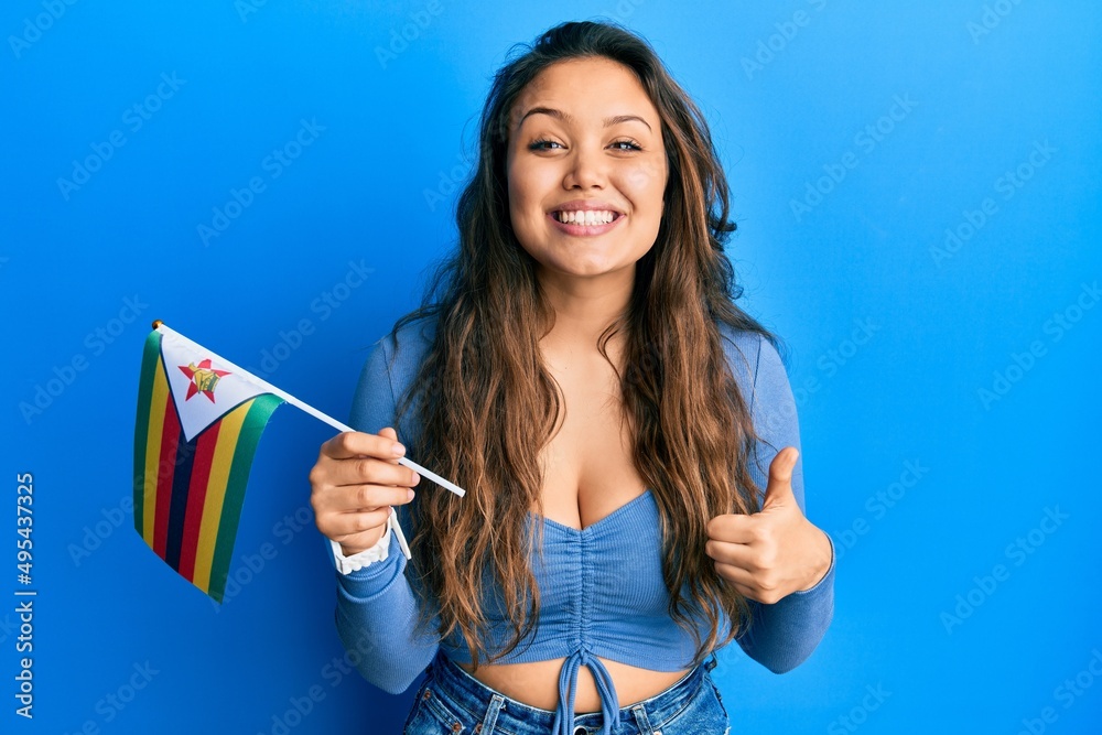 Young hispanic girl holding zimbabwe flag smiling happy and positive, thumb up doing excellent and approval sign Young hispanic girl holding zimbabwe flag smiling happy and positive, thumb up doing excellent and approval sign