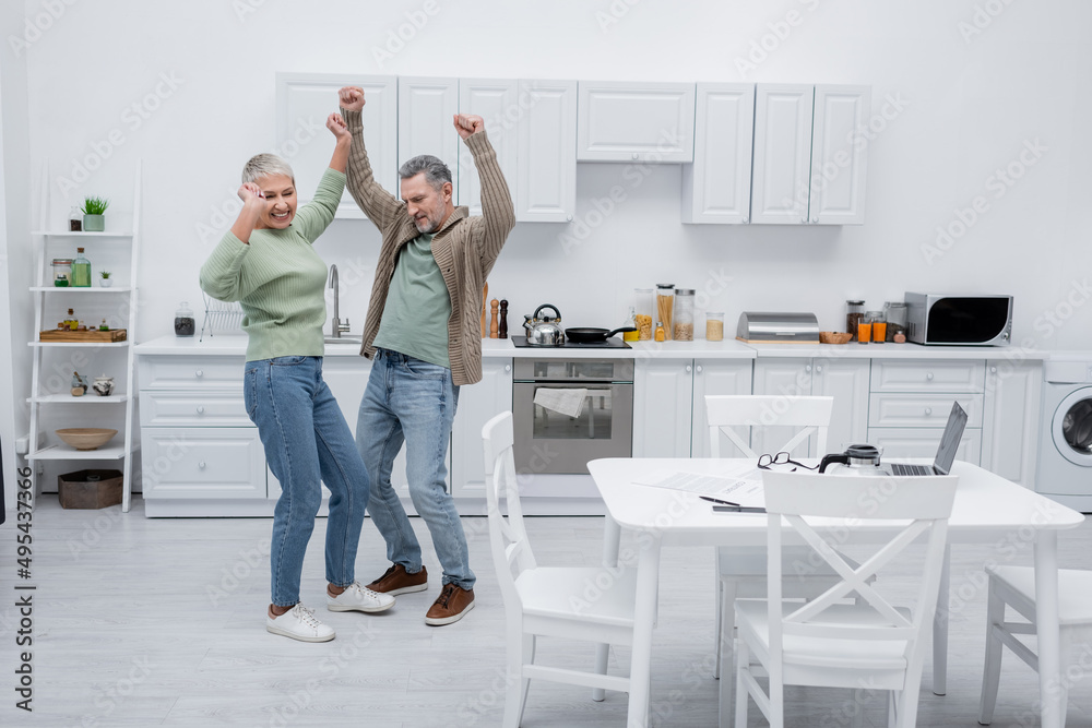 Happy couple dancing near papers and gadgets in kitchen.