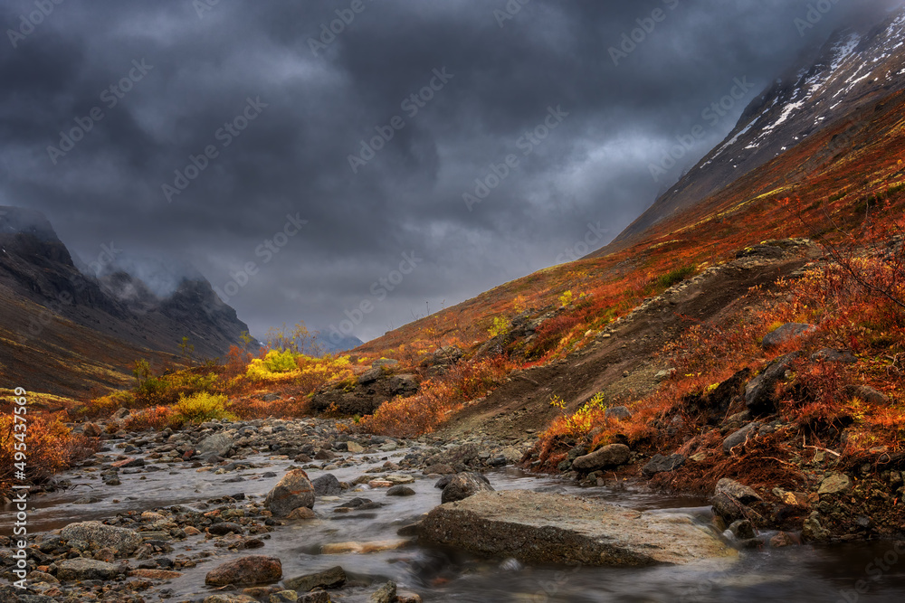 Stormy sky and fog in mountains. Autumn in tundra. Red and yellow autumn meadow landscape in Lapland with river and lake. 