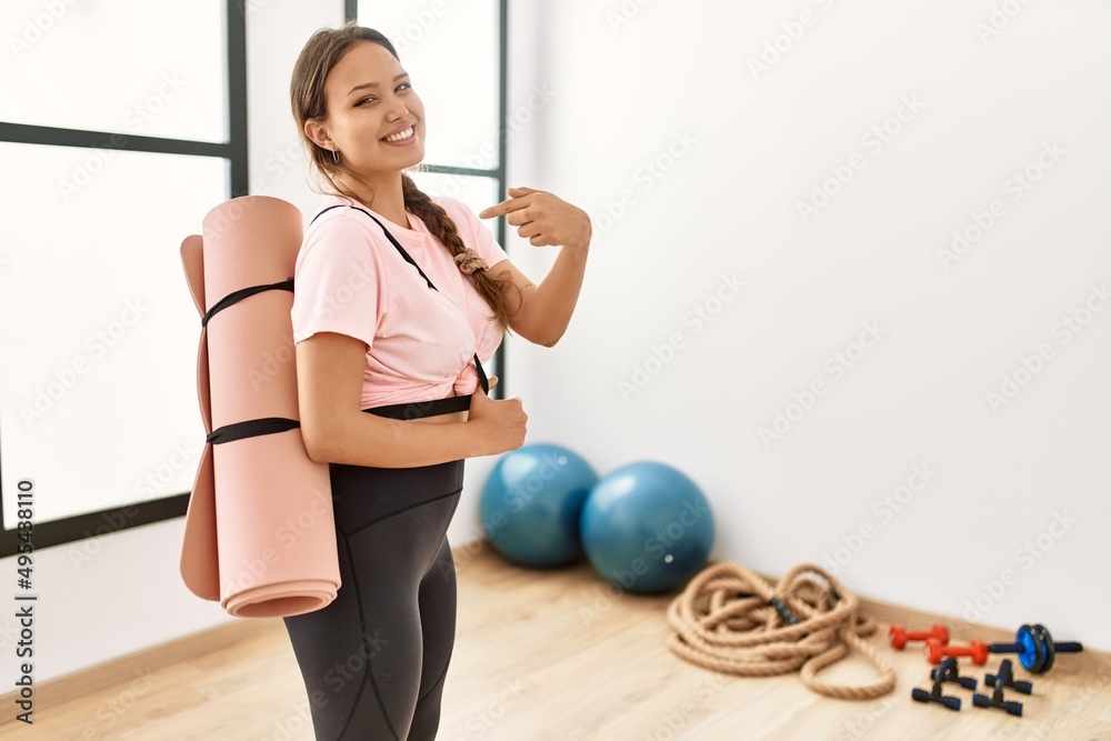 Young beautiful woman at the gym wearing sportswear holding yoga mat smiling happy pointing with hand and finger Young beautiful woman at the gym wearing sportswear holding yoga mat smiling happy pointing with hand and finger