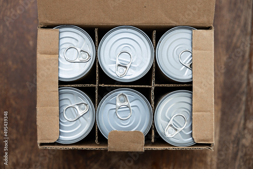Food cans in a open cardboard box on a wooden table. Concept image for supply chain disruption, food shortage, stockpiling in times of war.