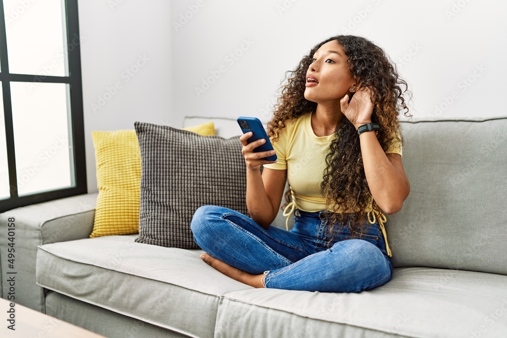 Beautiful hispanic woman sitting on the sofa at home using smartphone smiling with hand over ear listening an hearing to rumor or gossip. deafness concept. Beautiful hispanic woman sitting on the sofa at home using smartphone smiling with hand over ear listening an hearing to rumor or gossip. deafness concept.