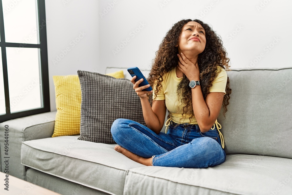 Beautiful hispanic woman sitting on the sofa at home using smartphone touching painful neck, sore throat for flu, clod and infection Beautiful hispanic woman sitting on the sofa at home using smartphone touching painful neck, sore throat for flu, clod and infection