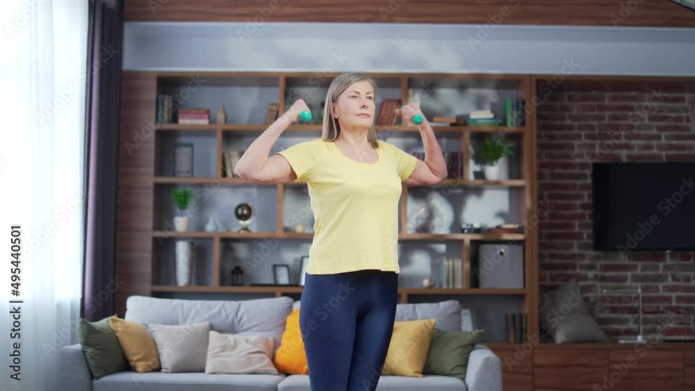 Sporty Old senior gray-haired woman exercising while standing with dumbbells in her hands in a room at home. Older Mature female doing fitness workout indoors. Concept about people, gym at home, sport Sporty Old senior gray-haired woman exercising while standing with dumbbells in her hands in a room at home. Older Mature female doing fitness workout indoors. Concept about people, gym at home, sport