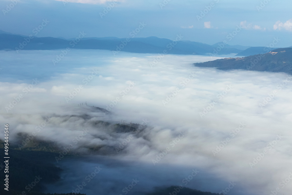 A bird's-eye view of the mountains shrouded in thick fog. In the background are dark silhouettes of ridges and a cloudy sky.