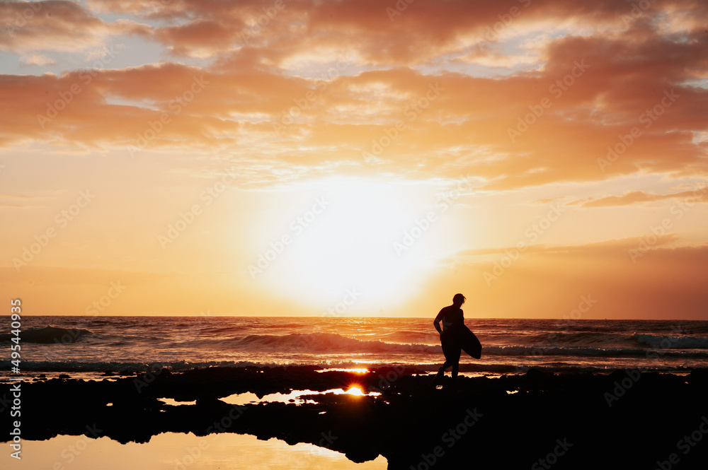 Silhouette of unrecognizable surfer walking with his surfboard with beautiful dramatic sky during sunset on background.