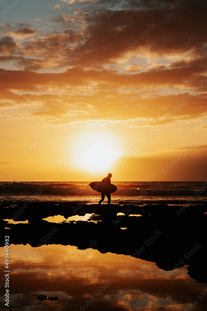 Vertical shot silhouette of unrecognizable male surfer walking with his surfboard on the rocks. Dramatic red and orange sky during sunset and reflection of the waves.