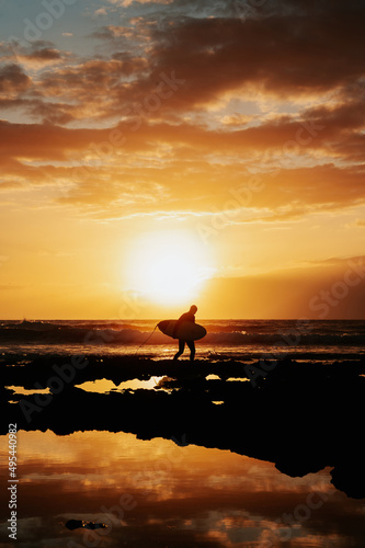 Vertical shot silhouette of unrecognizable male surfer walking with his surfboard on the rocks. Dramatic red and orange sky during sunset and reflection of the waves. © Pintau Studio