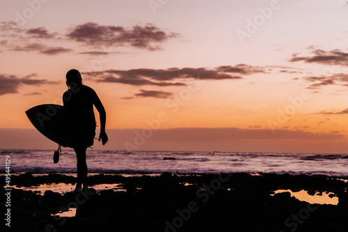 Unrecognizable silhouette of male surfer with his surfboard walking away. Backlight during sunset with dramatic sky and reflection.