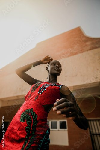 Low angle view of young African woman in a red dress standing beside building photo