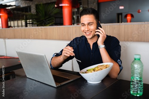 Handsome businessperson talking at the phone and using computer during his lunch break while eating salad.