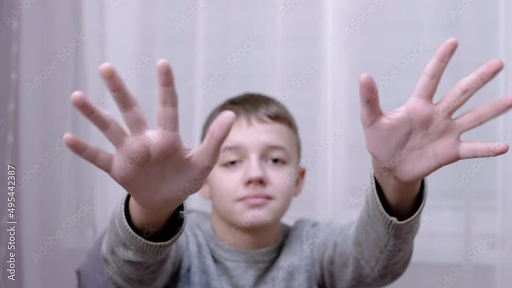 Sad Child Waving Hands Goodbye, Hello, Bye while Sitting on an Armchair in Room. Close-up. A teenager is gesturing with hands. Boy is upset because of the departure of the guests. Blurred background. Sad Child Waving Hands Goodbye, Hello, Bye while Sitting on an Armchair in Room. Close-up. A teenager is gesturing with hands. Boy is upset because of the departure of the guests. Blurred background.