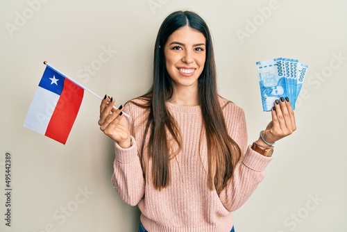 Fotografi Young hispanic woman holding chile flag and chilean pesos banknotes smiling with a happy and cool smile on face