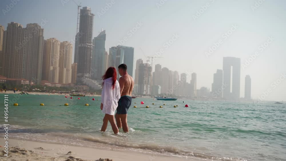 Married couple walks along a beautiful sea beach in Dubai against the backdrop of the cityscape and skyscrapers. Honeymoon in the United Arab Emirates. Travel and recreation. 