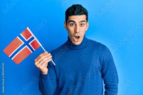 Handsome hispanic man holding norway flag scared and amazed with open mouth for surprise, disbelief face