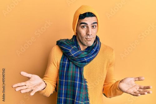 Handsome hispanic man wearing wool hat and winter scarf clueless and confused expression with arms and hands raised. doubt concept. © Krakenimages.com