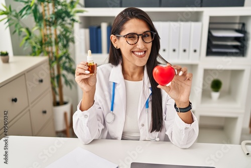 Young hispanic doctor woman holding heart and cbd oil winking looking at the camera with sexy expression, cheerful and happy face.