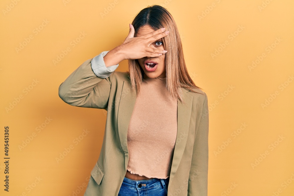 Beautiful hispanic woman wearing business jacket peeking in shock covering face and eyes with hand, looking through fingers with embarrassed expression.