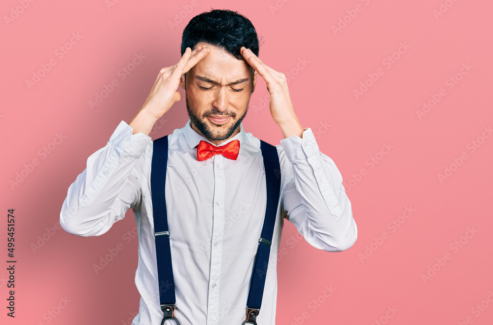 Hispanic man with beard wearing hipster look with bow tie and suspenders with hand on head for pain in head because stress. suffering migraine. Hispanic man with beard wearing hipster look with bow tie and suspenders with hand on head for pain in head because stress. suffering migraine.