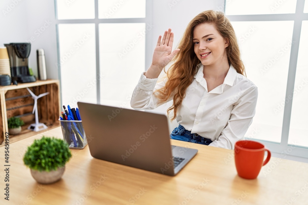 Young caucasian woman working at the office using computer laptop waiving saying hello happy and smiling, friendly welcome gesture Young caucasian woman working at the office using computer laptop waiving saying hello happy and smiling, friendly welcome gesture