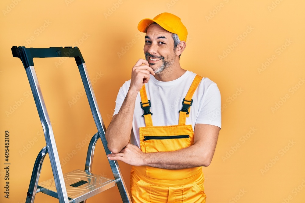 Handsome middle age man with grey hair holding ladder looking confident at the camera with smile with crossed arms and hand raised on chin. thinking positive. Handsome middle age man with grey hair holding ladder looking confident at the camera with smile with crossed arms and hand raised on chin. thinking positive.