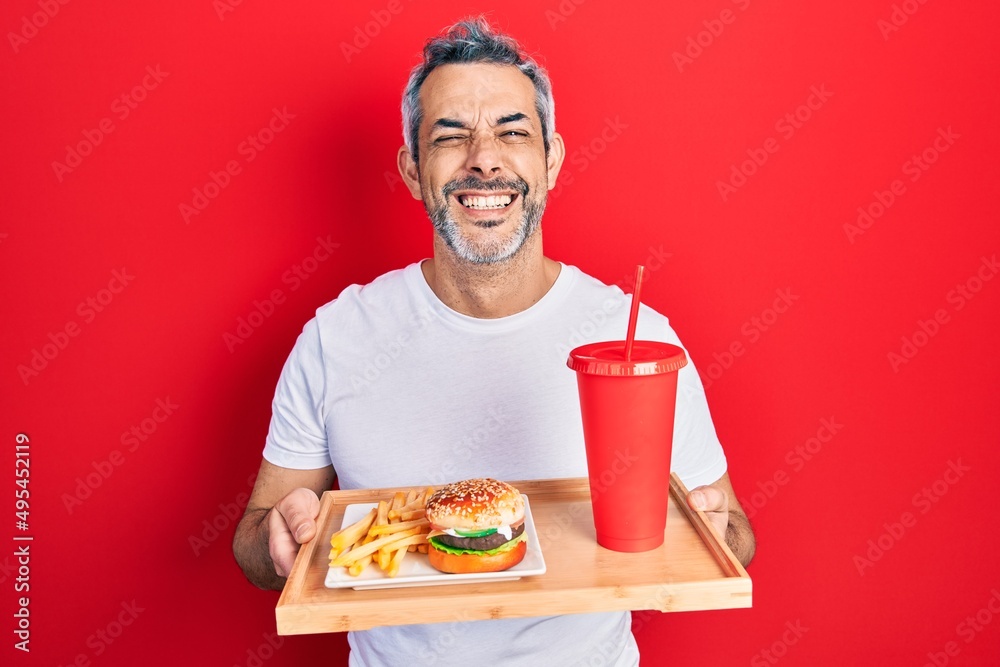 Handsome middle age man with grey hair eating a tasty classic burger with fries and soda smiling and laughing hard out loud because funny crazy joke. Handsome middle age man with grey hair eating a tasty classic burger with fries and soda smiling and laughing hard out loud because funny crazy joke.