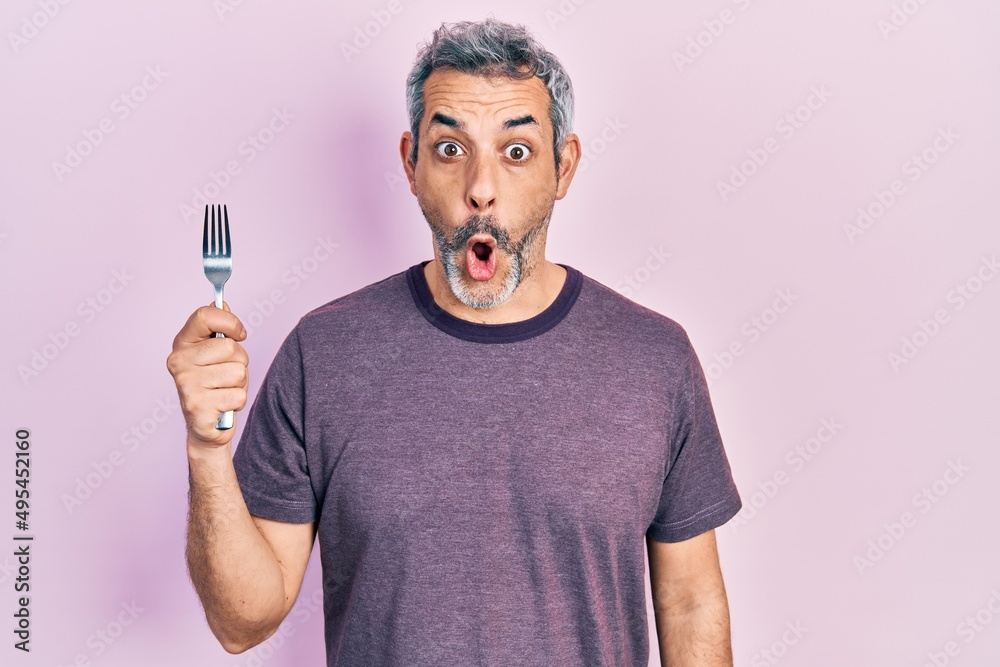 Handsome middle age man with grey hair holding one silver fork scared and amazed with open mouth for surprise, disbelief face Handsome middle age man with grey hair holding one silver fork scared and amazed with open mouth for surprise, disbelief face