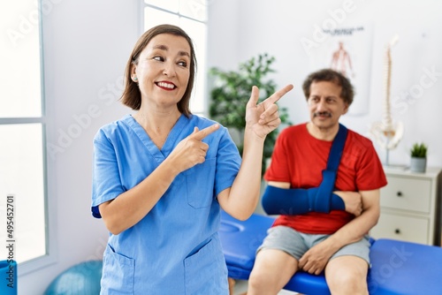 Middle age doctor woman with patient with arm injury at rehabilitation clinic smiling and looking at the camera pointing with two hands and fingers to the side.