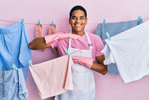Young handsome hispanic man wearing cleaner apron holding clothes on clothesline gesturing with hands showing big and large size sign, measure symbol. smiling looking at the camera. measuring concept.