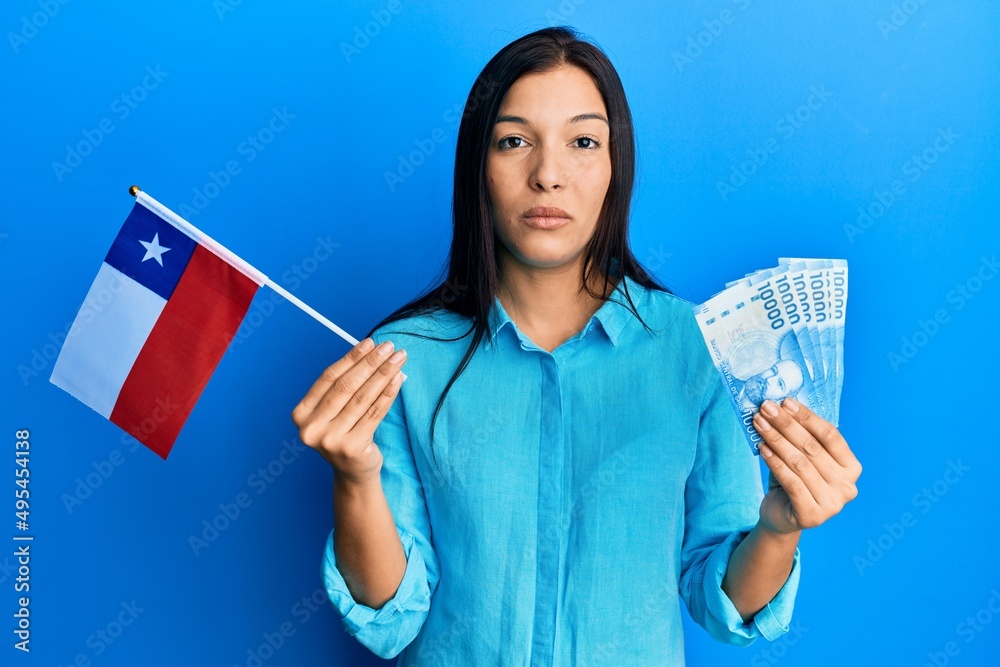 Young latin woman holding chile flag and chilean pesos banknotes relaxed with serious expression on face. simple and natural looking at the camera. Young latin woman holding chile flag and chilean pesos banknotes relaxed with serious expression on face. simple and natural looking at the camera.