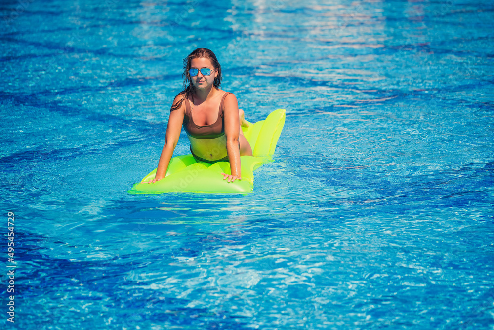 Young sexy woman in a swimsuit swims in the pool on an inflatable mattress. Girl in sunglasses with a smile on faces in a blue pool on vacation