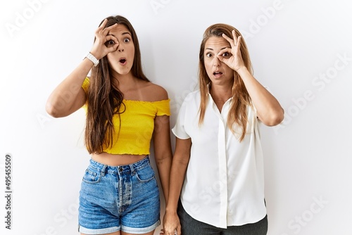 Mother and daughter together standing together over isolated background doing ok gesture shocked with surprised face, eye looking through fingers. unbelieving expression.