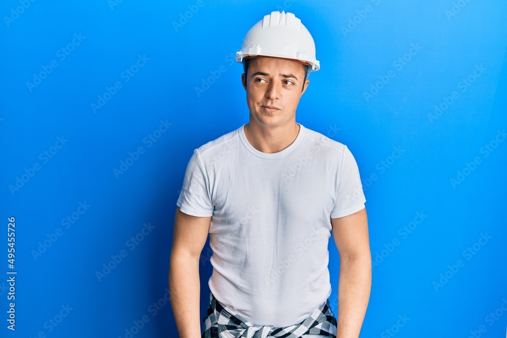 Handsome young man wearing builder uniform and hardhat smiling looking to the side and staring away thinking.