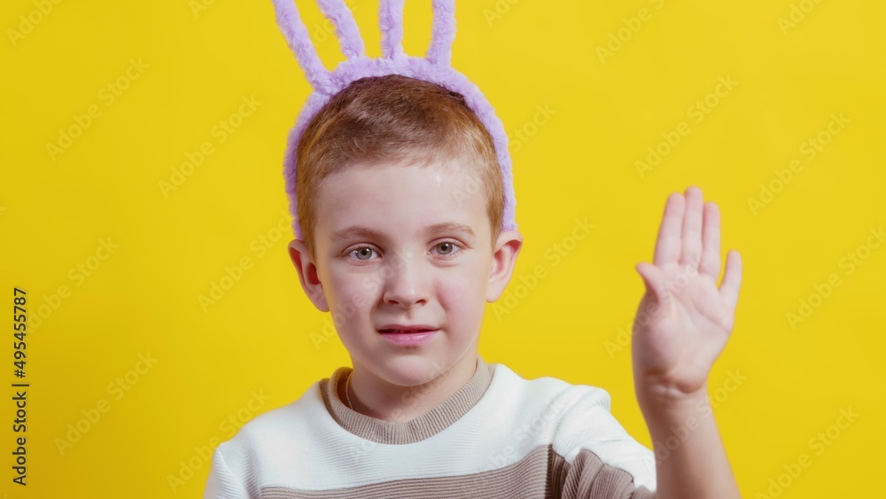 Close-up of a happy boy with rabbit ears on a yellow background. The boy waves his hand and looks at the camera. Easter Close-up of a happy boy with rabbit ears on a yellow background. The boy waves his hand and looks at the camera. Easter