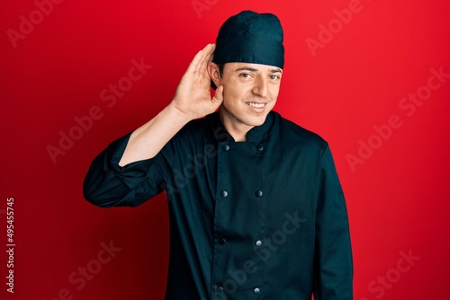 Handsome young man wearing professional cook uniform and hat smiling with hand over ear listening an hearing to rumor or gossip. deafness concept.