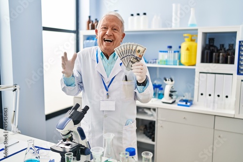 Senior scientist with grey hair working at laboratory holding dollars celebrating victory with happy smile and winner expression with raised hands
