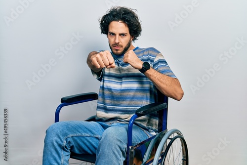 Handsome hispanic man sitting on wheelchair punching fist to fight, aggressive and angry attack, threat and violence