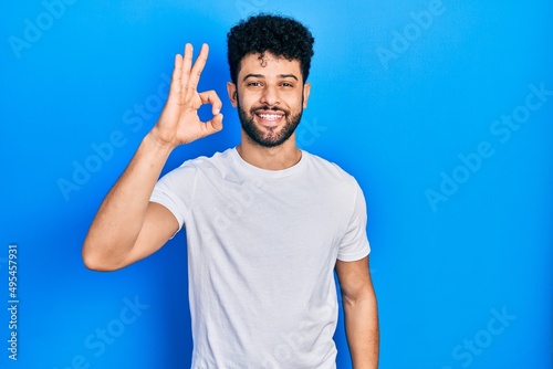 Young arab man with beard wearing casual white t shirt smiling positive doing ok sign with hand and fingers. successful expression.