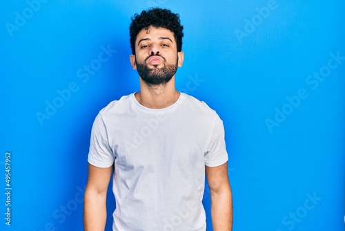 Young arab man with beard wearing casual white t shirt looking at the camera blowing a kiss on air being lovely and sexy. love expression.