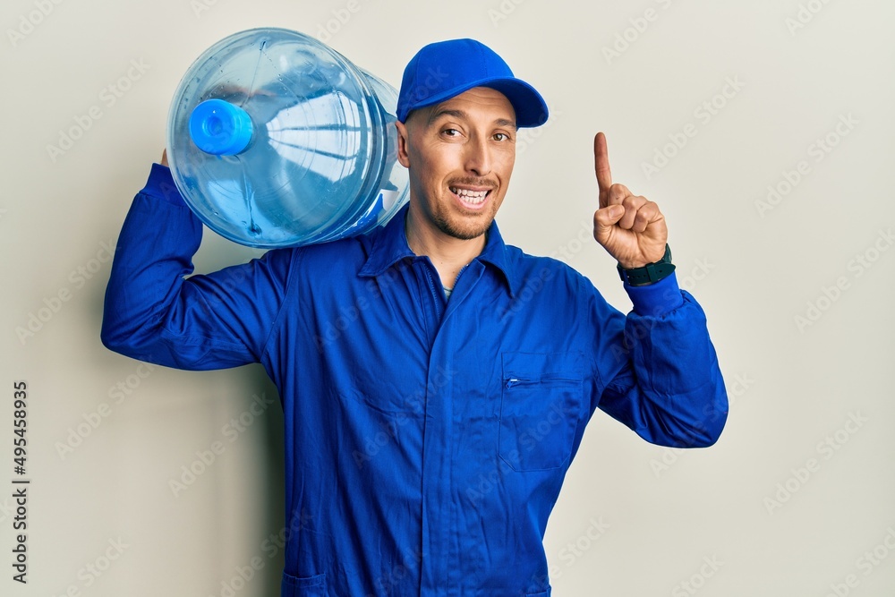 Bald courier man with beard holding a gallon bottle of water for delivery surprised with an idea or question pointing finger with happy face, number one