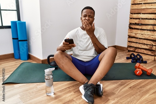 Young african man sitting on training mat at the gym using smartphone bored yawning tired covering mouth with hand. restless and sleepiness.