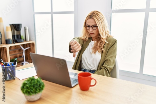 Beautiful blonde woman working at the office with laptop looking unhappy and angry showing rejection and negative with thumbs down gesture. bad expression.