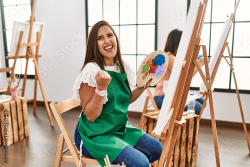 Young hispanic artist women painting on canvas at art studio very happy and excited doing winner gesture with arms raised, smiling and screaming for success. celebration concept.