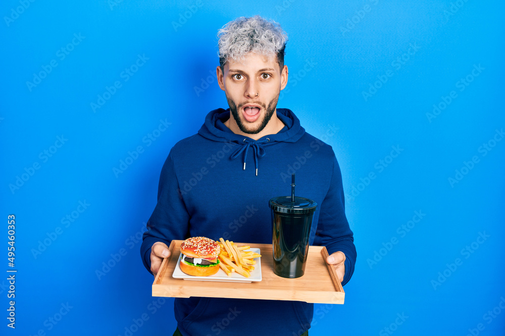 Young hispanic man with modern dyed hair eating a tasty classic burger with fries and soda afraid and shocked with surprise and amazed expression, fear and excited face. Young hispanic man with modern dyed hair eating a tasty classic burger with fries and soda afraid and shocked with surprise and amazed expression, fear and excited face.